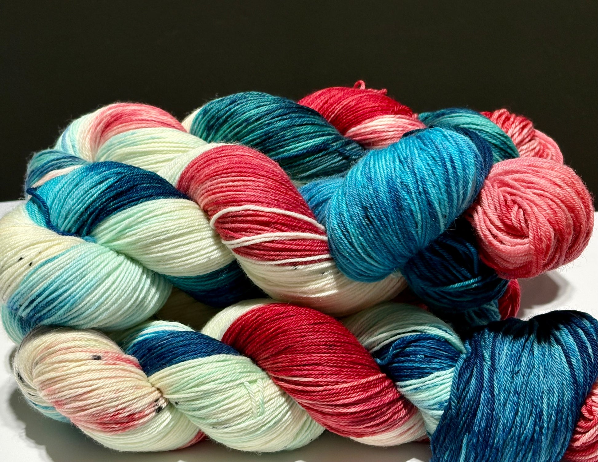 Multiple skeins of Dawn’s Early Light hand-dyed yarn stacked together, showing bold stripes of deep navy, aqua, creamy white, and vermilion red. Each skein has unique color placement while maintaining the same patriotic palette.