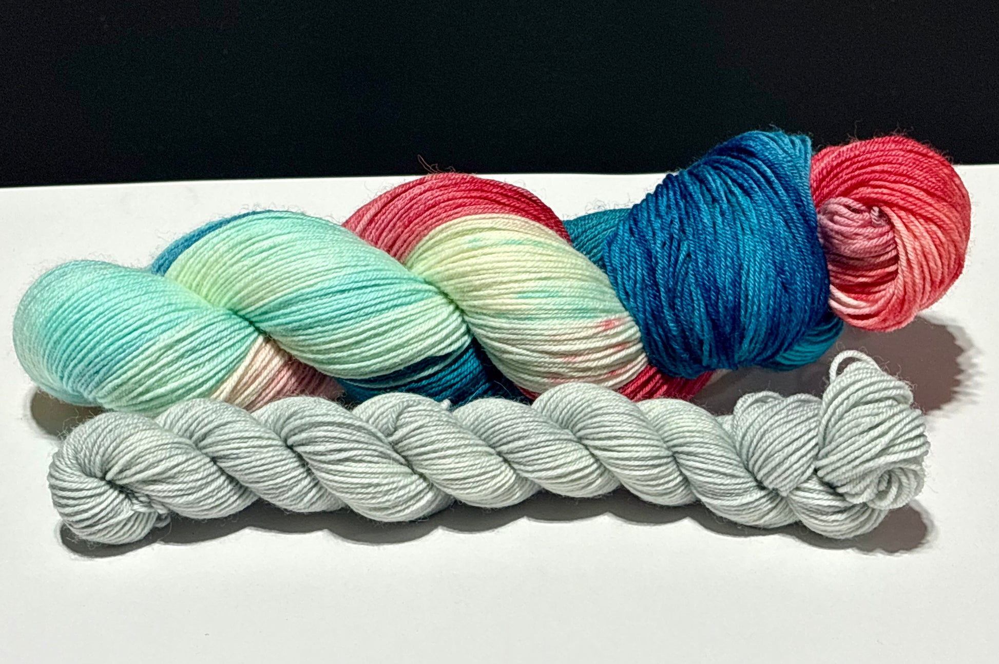 Close-up of Dawn’s Early Light hand-dyed yarn highlighting saturated navy blue, aqua, creamy white, and vermilion red with scattered charcoal speckles. The coordinating pale gray mini skein complements the sock set.
