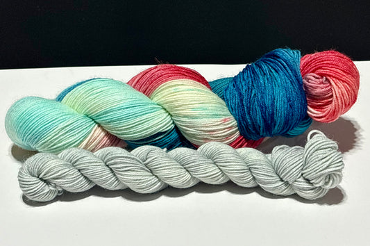 Close-up of Dawn’s Early Light hand-dyed yarn highlighting saturated navy blue, aqua, creamy white, and vermilion red with scattered charcoal speckles. The coordinating pale gray mini skein complements the sock set.